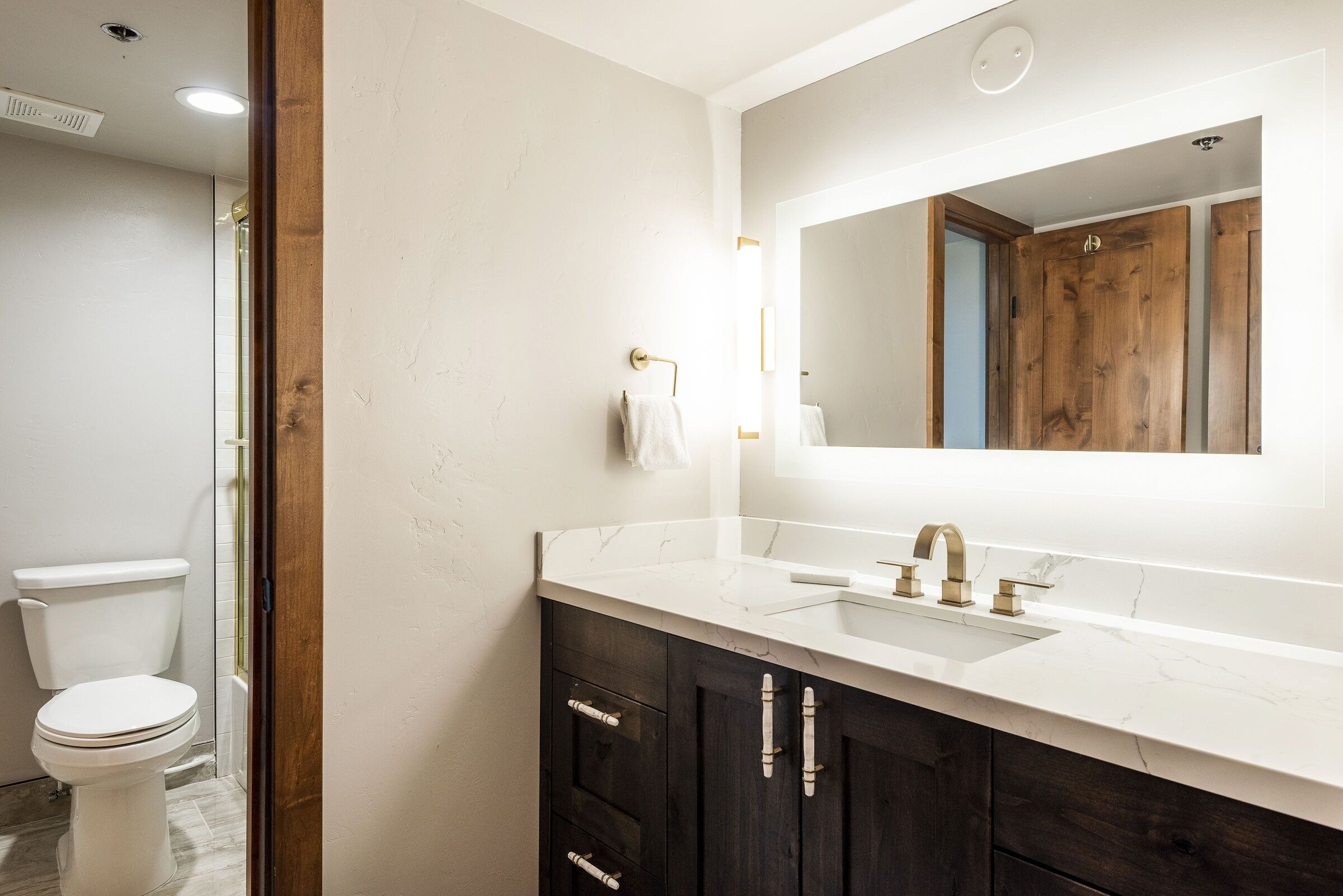 A bathroom with a white toilet, dark wooden cabinets, a white sink with a gold faucet, and a large mirror.