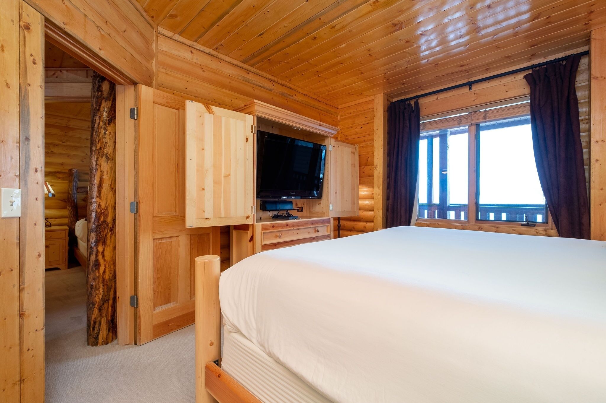 A wooden cabin room with a bed, a TV, and a window with curtains.