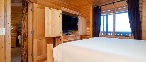 A wooden cabin room with a bed, a TV, and a window with curtains.