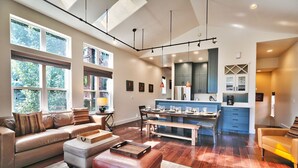 A spacious living area with a large sofa, ottoman, and dining table. There is a kitchen area with blue cabinets and a refrigerator. - Woodside Haus 3 Bedroom Home by RedAwning (Park City)