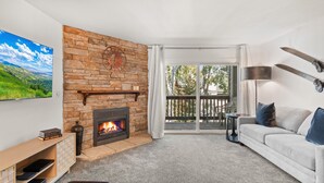 A living room with a stone fireplace, a large TV, a sofa, and a balcony with a view of greenery.