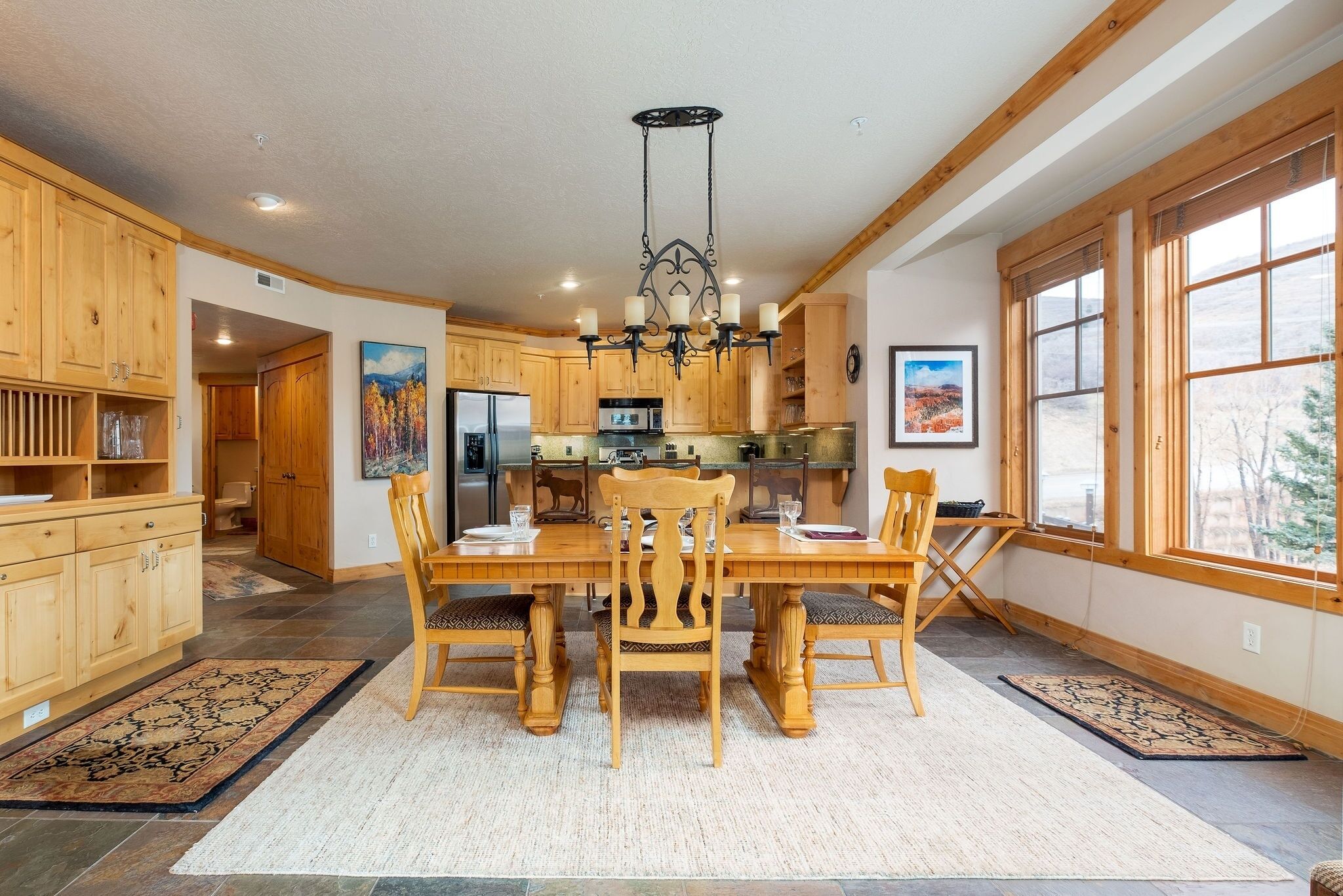 A spacious kitchen with wooden cabinets, a dining table with chairs, a chandelier, and large windows.