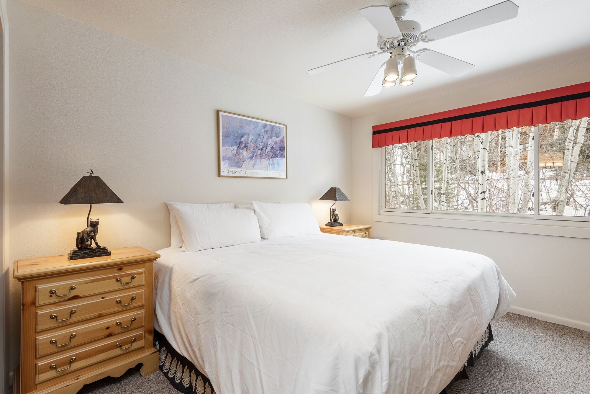 A bedroom with a bed, two nightstands, a ceiling fan, a window with a view of trees, and a painting on the wall.