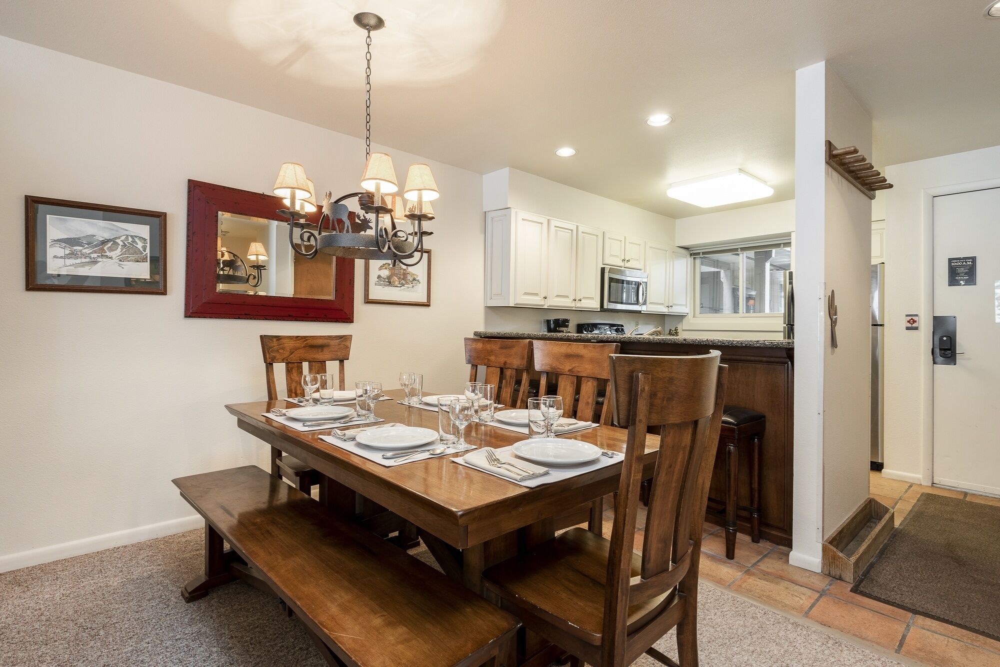 Un coin repas avec une table en bois dressée pour six personnes, un lustre et des meubles de cuisine en arrière-plan.