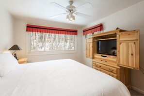 A bedroom with a bed, a wooden TV cabinet, a ceiling fan, and a window with a view of trees. - Snow Flower 145 2 Bedroom Condo by RedAwning (Park City)
