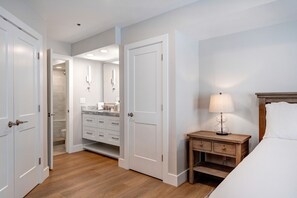 A hotel room with a wooden floor, a bed with white linens, a nightstand with a lamp, a wooden dresser, and a bathroom with a shower and toilet visible through an open door. - Silver Cliff Village 103 2 Bedroom Condo by RedAwning (Park City)
