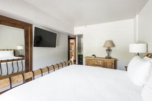 A bedroom with a large bed, wooden headboard, a TV mounted on the wall, a wooden dresser, and a lamp.