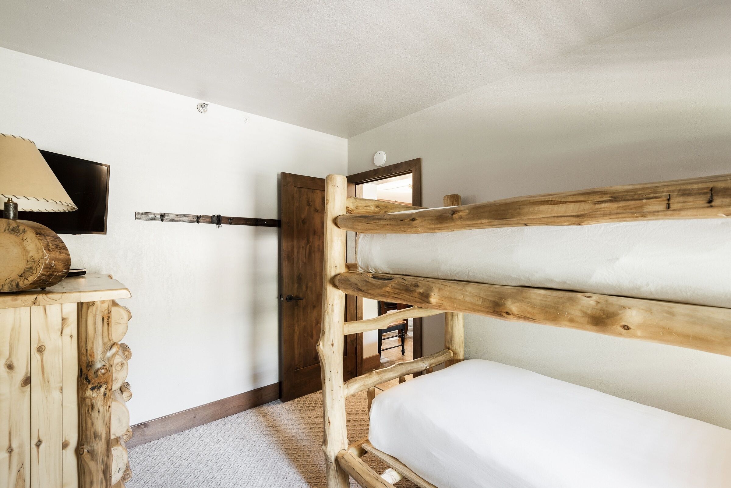 A bunk bed with a wooden headboard, a wooden dresser with a lamp, and a television on the wall.