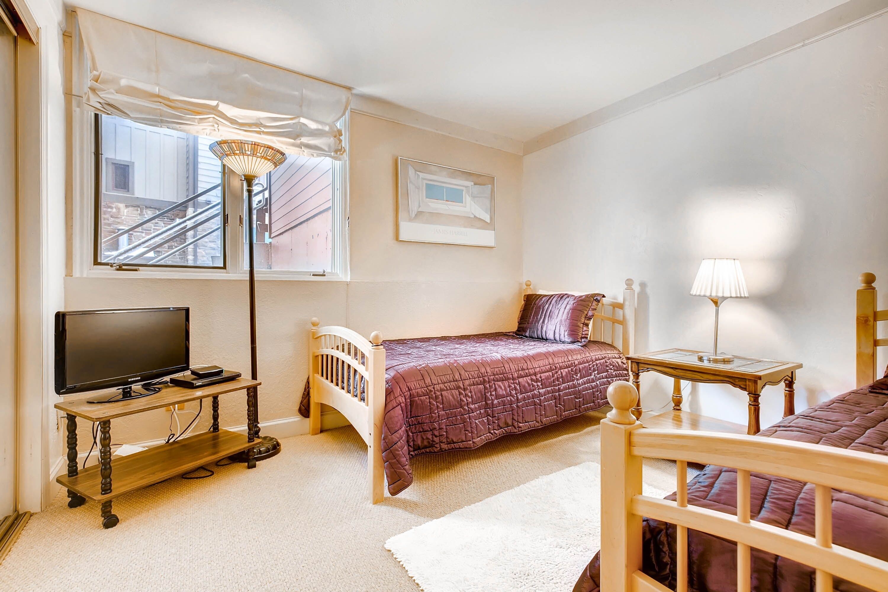 A bedroom with a bed, a TV on a wooden stand, a lamp, and a window with curtains.