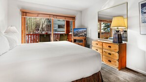 A hotel room with a large bed, a desk, a chair, a dresser, a mirror, and a window with a view of trees and buildings. - Snow Flower 065 2 Bedroom Condo by RedAwning (Park City)