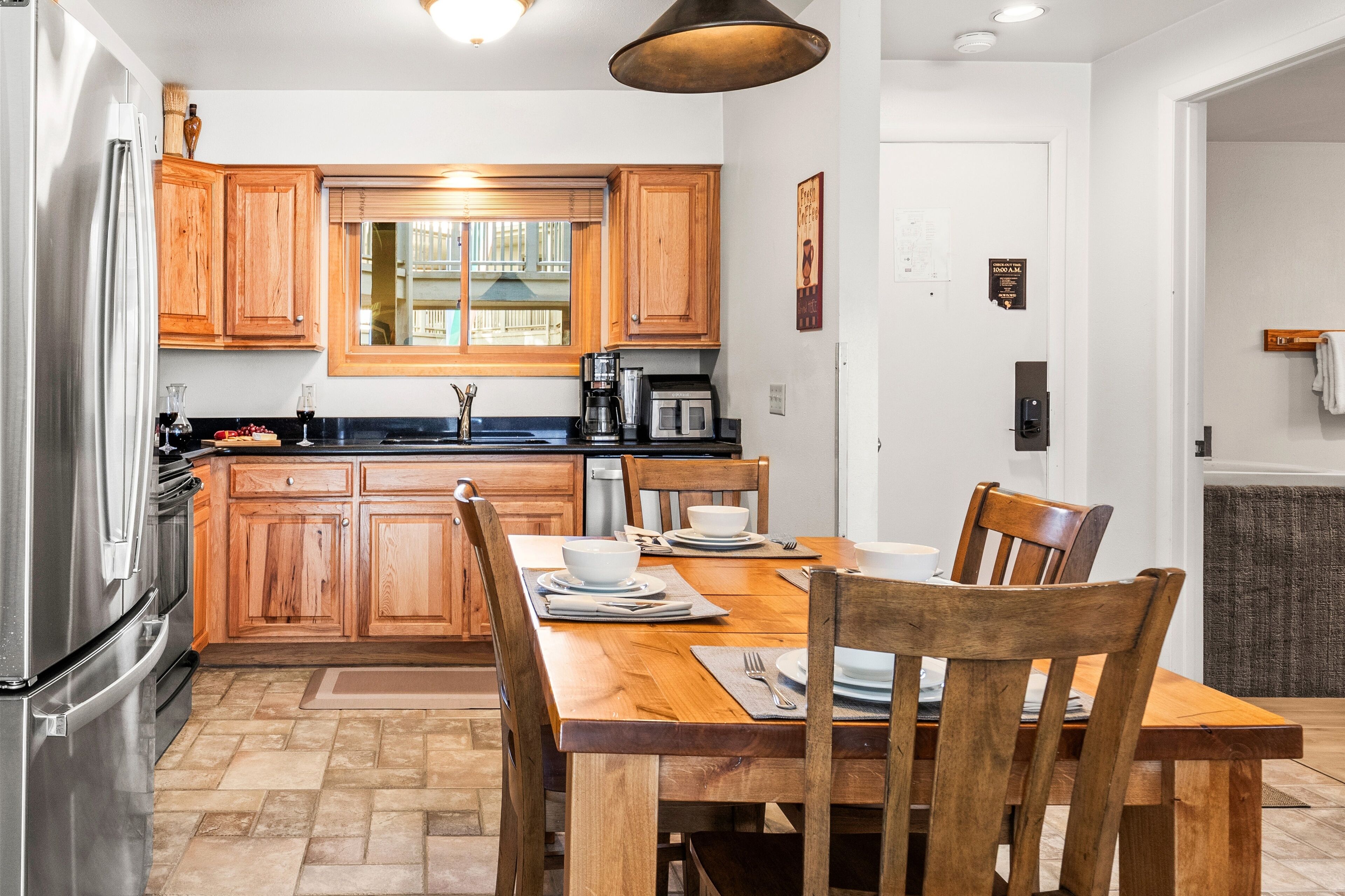 A kitchen with wooden cabinets, a stainless steel refrigerator, a dining table with chairs, and a window with blinds.