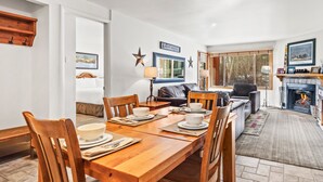 A dining area with a wooden table set for four, surrounded by chairs. There is a fireplace with a mantel and framed pictures above it. - Snow Flower 065 2 Bedroom Condo by RedAwning (Park City)
