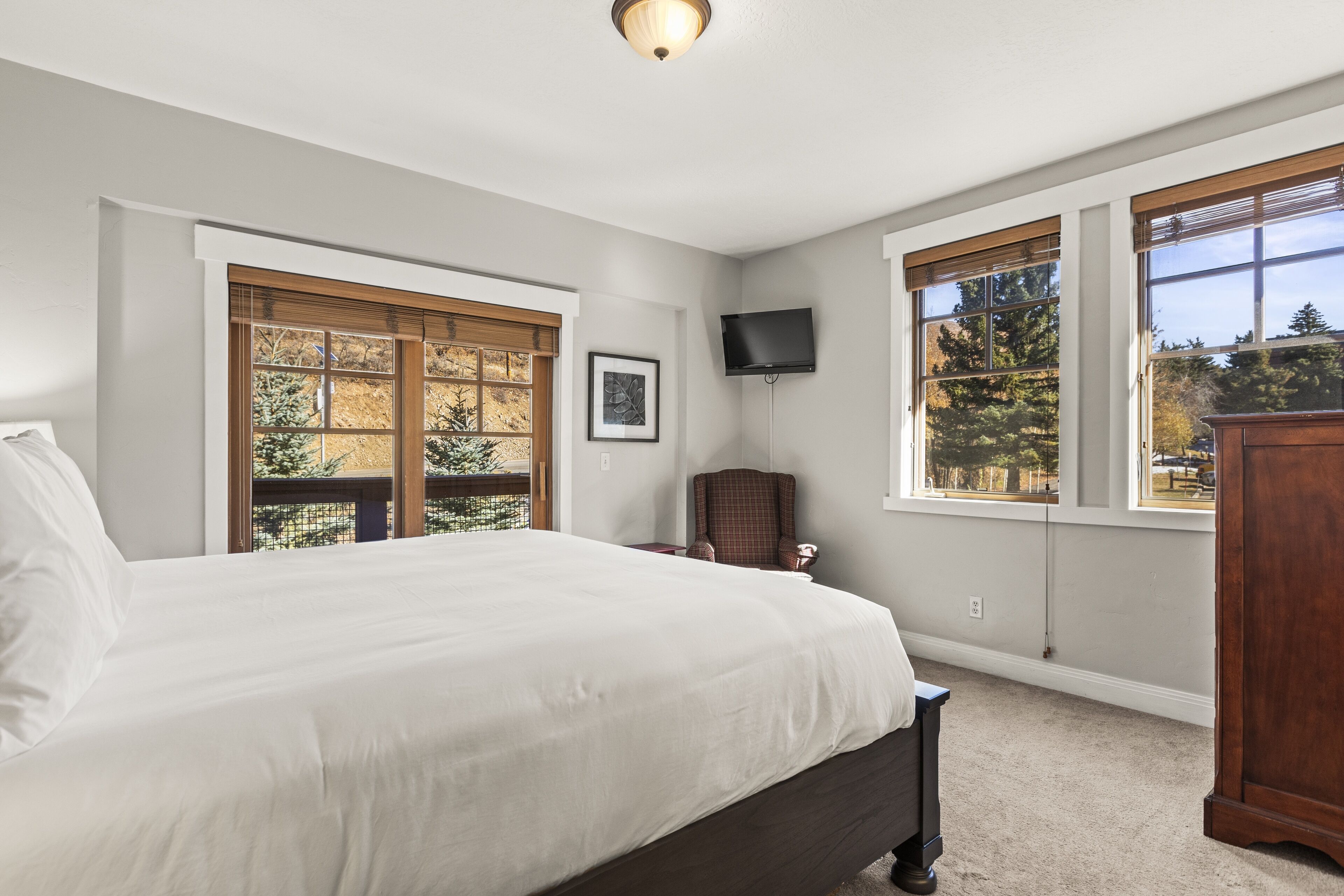 A bedroom with a large bed, two windows with blinds, a flat-screen TV mounted on the wall, and a wooden nightstand.