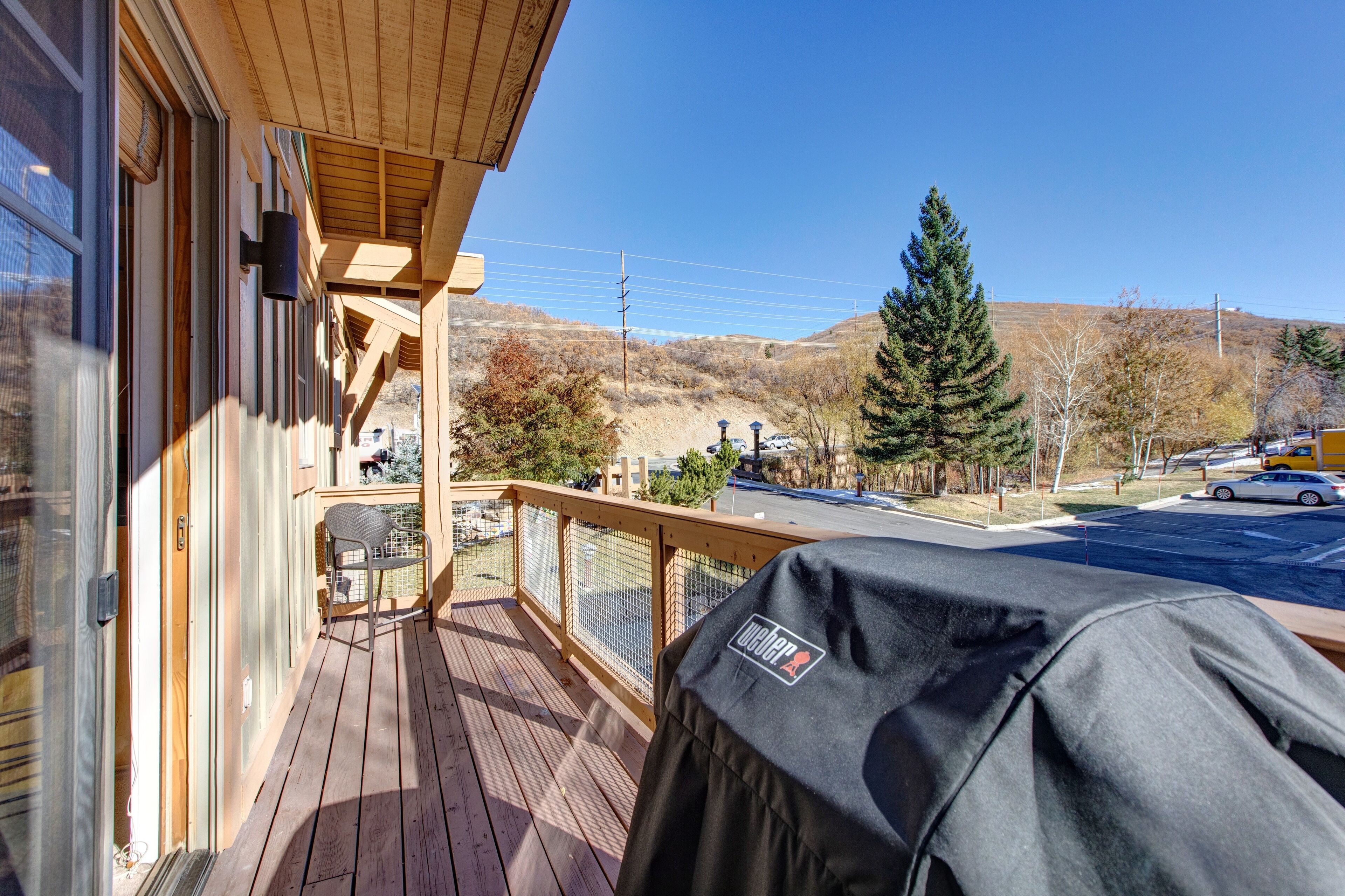 A wooden deck with a grill, a chair, and a view of a parking lot and hills.