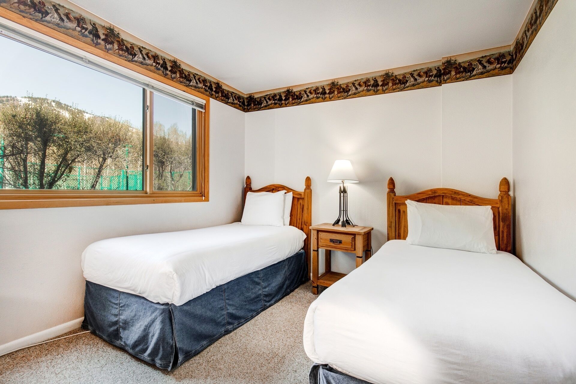 A bedroom with two single beds, a nightstand with a lamp, a window with a view of trees, and a decorative border on the ceiling.