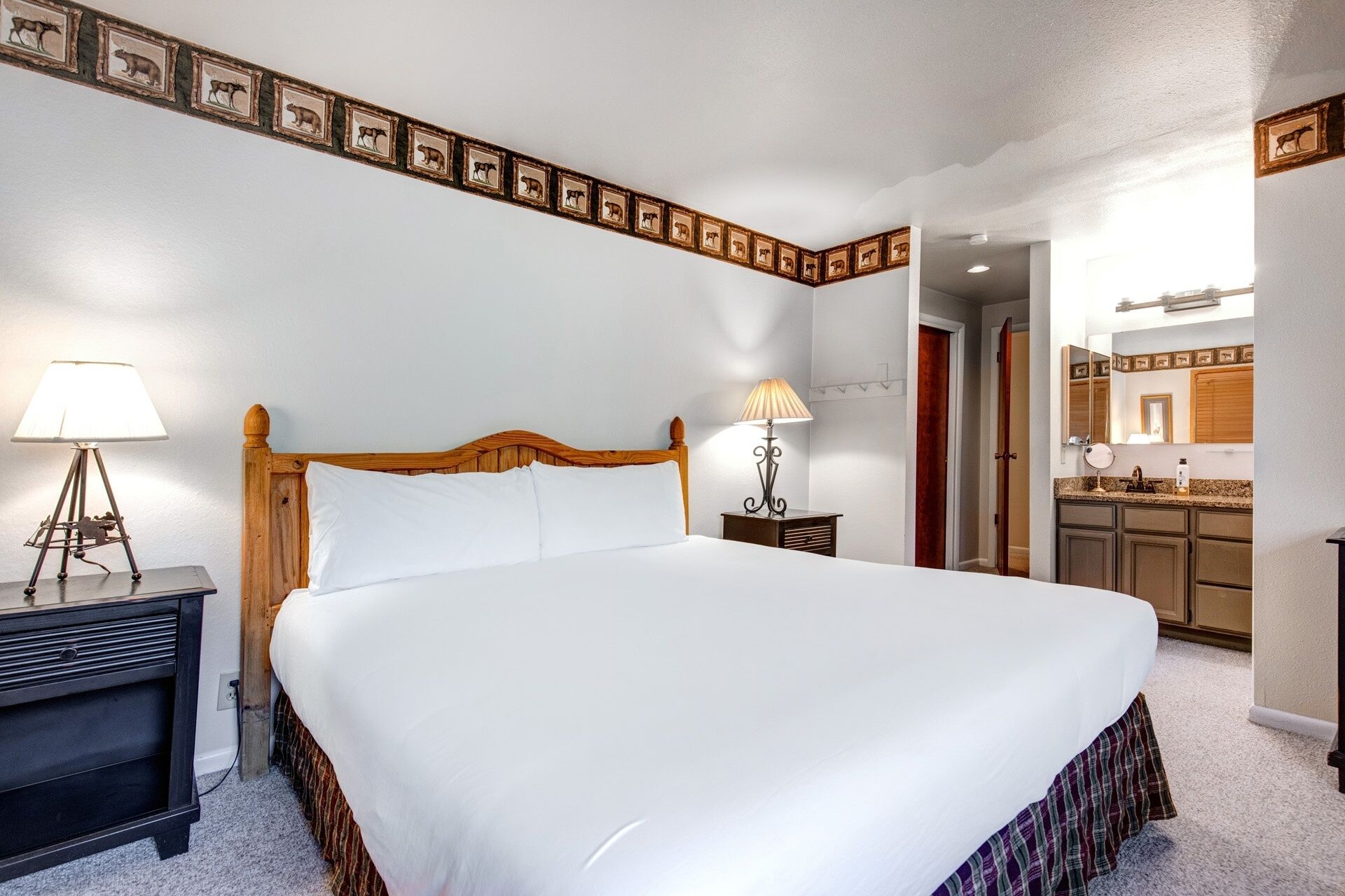 Habitación de hotel con una cama grande, lámparas de noche, cabecero de madera, una mesita pequeña, un espejo y una kitchenette.