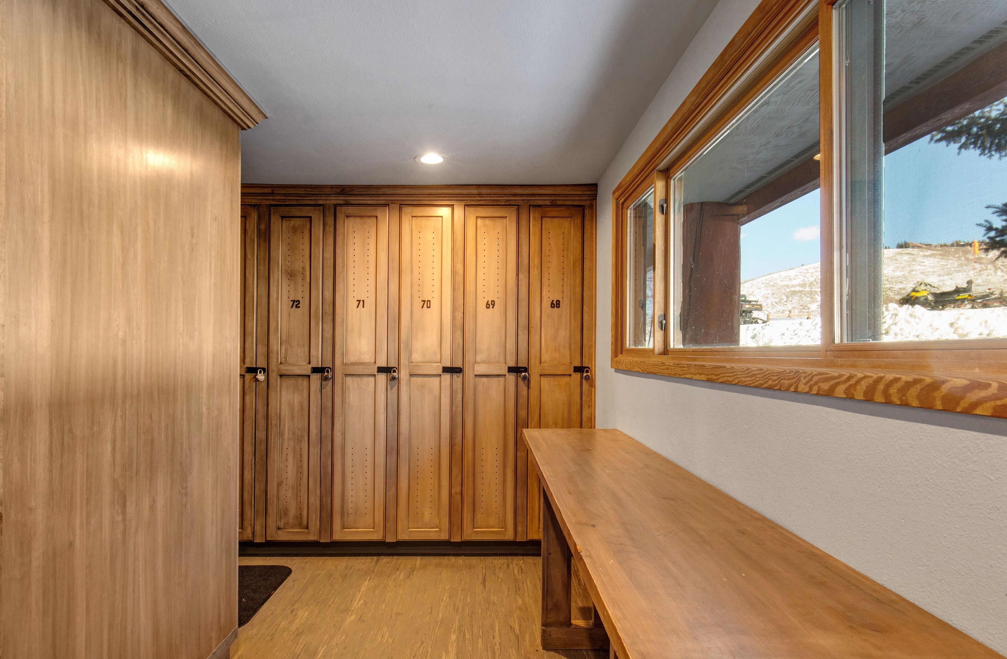A wooden locker room with numbered lockers, a bench, and a window with a view of a snowy landscape.