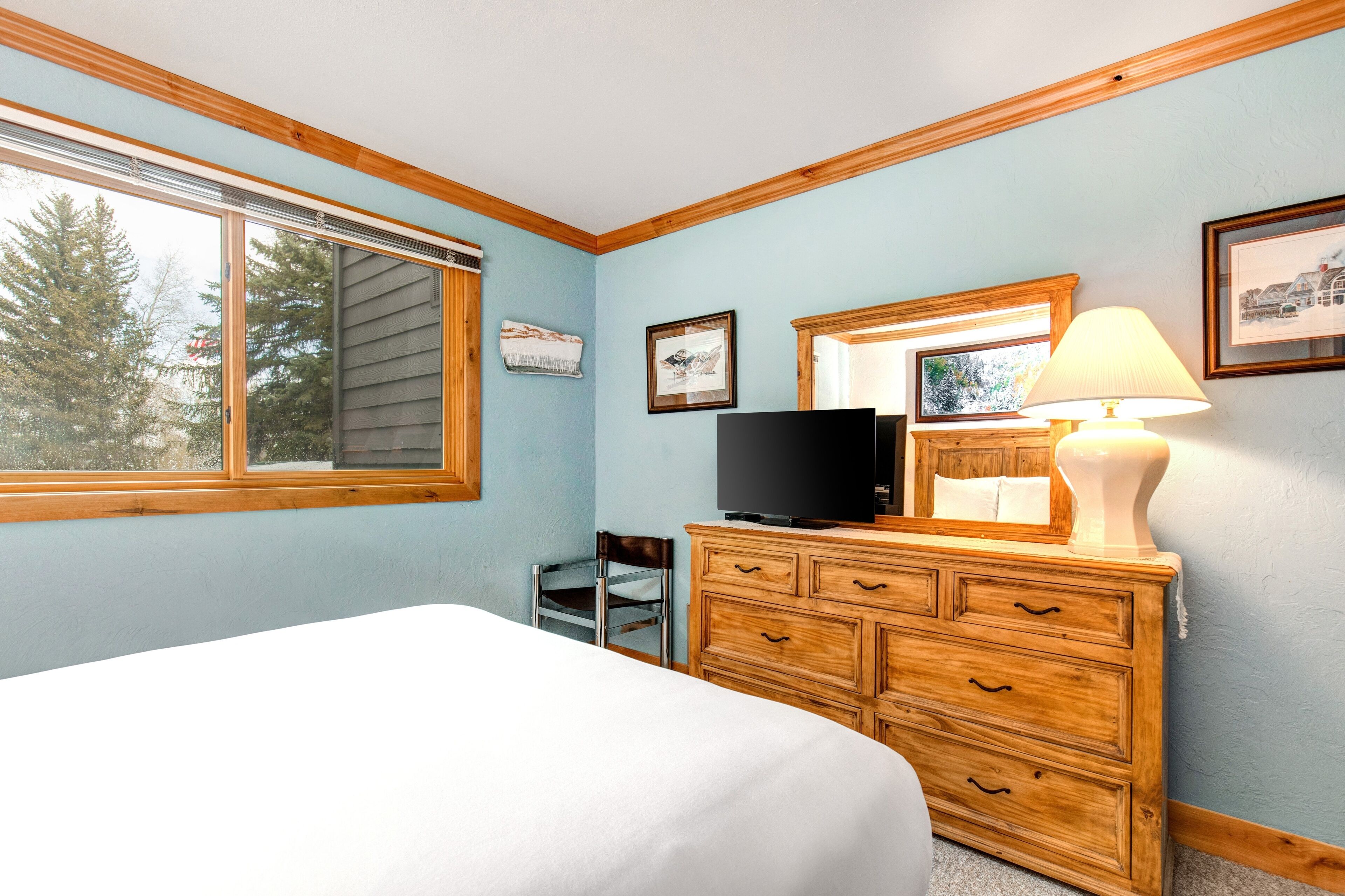 A bedroom with a bed, wooden dresser, television, and a window with a view of trees.