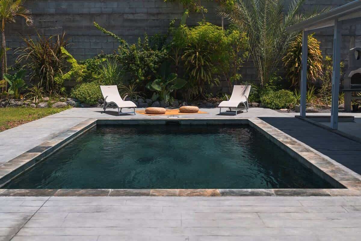 Una piscina rectangular con dos sillas reclinables blancas y un muro de piedra al fondo.