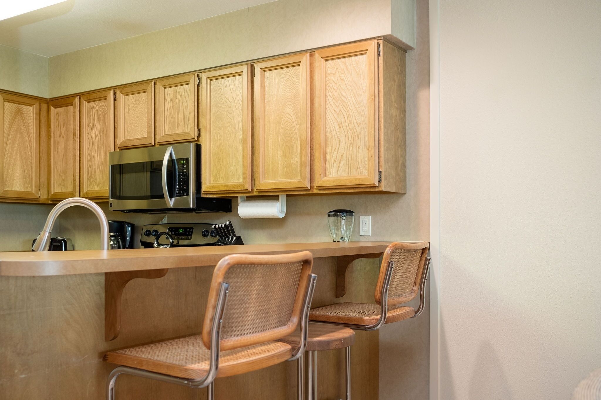 A kitchen with wooden cabinets, a stainless steel microwave, a sink, and two wicker bar stools.