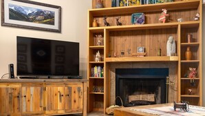 A living room with a television, wooden shelves, a fireplace, and a table. - Snow Flower 159 2 Bedroom Condo by RedAwning (Park City)