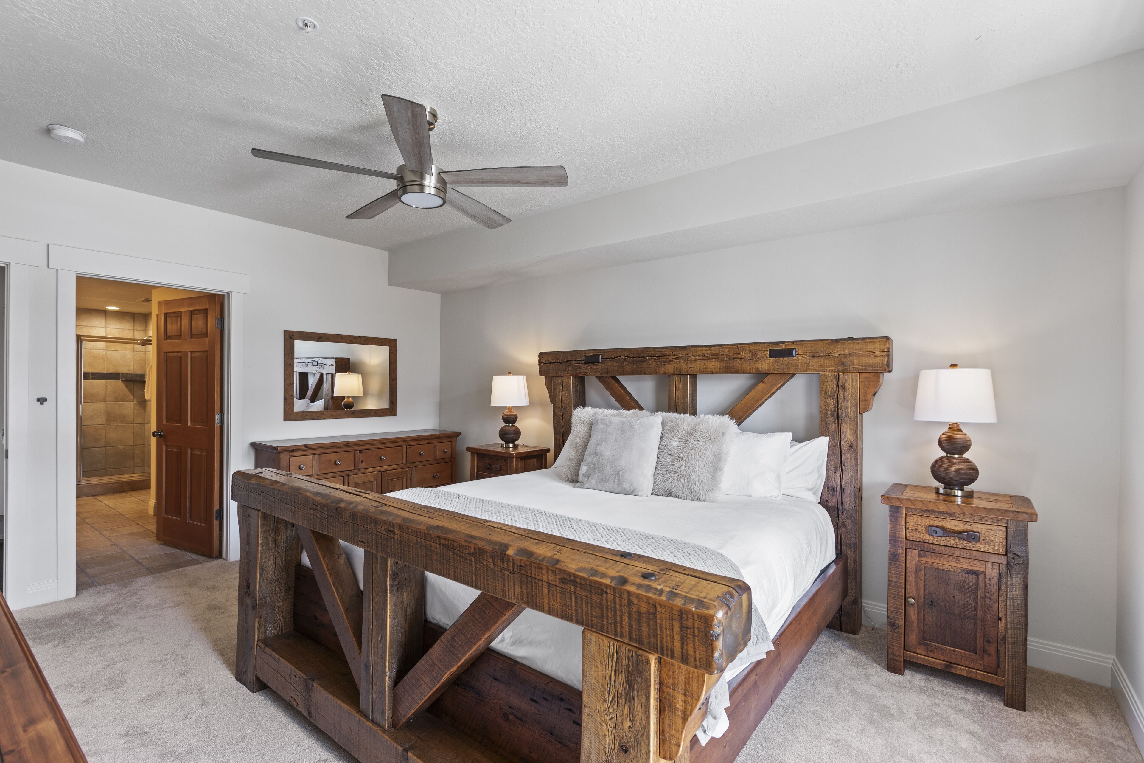 A bedroom with a wooden bed frame, two bedside tables with lamps, a ceiling fan, and a door leading to another room.