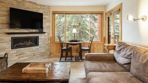 A cozy living room with a stone fireplace, a flat-screen TV, a wooden dining table with chairs, and a large window offering a view of trees. - Snow Flower 055 2 Bedroom Condo by RedAwning (Park City)