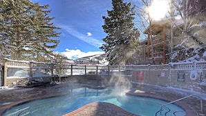 A hot tub with a brick border, surrounded by snow-covered trees and a building with a balcony. - Snow Flower 055 2 Bedroom Condo by RedAwning (Park City)