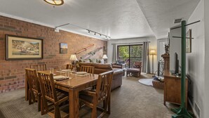 A dining area with a brick wall, wooden table, and chairs. There is a staircase with railings and a window with curtains. - Park Station 121 2 Bedroom Condo by RedAwning (Park City)