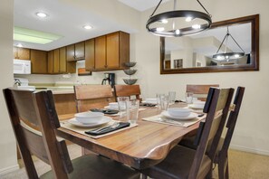 A dining area with a wooden table set for four, surrounded by chairs. There is a chandelier above the table and a mirror on the wall.