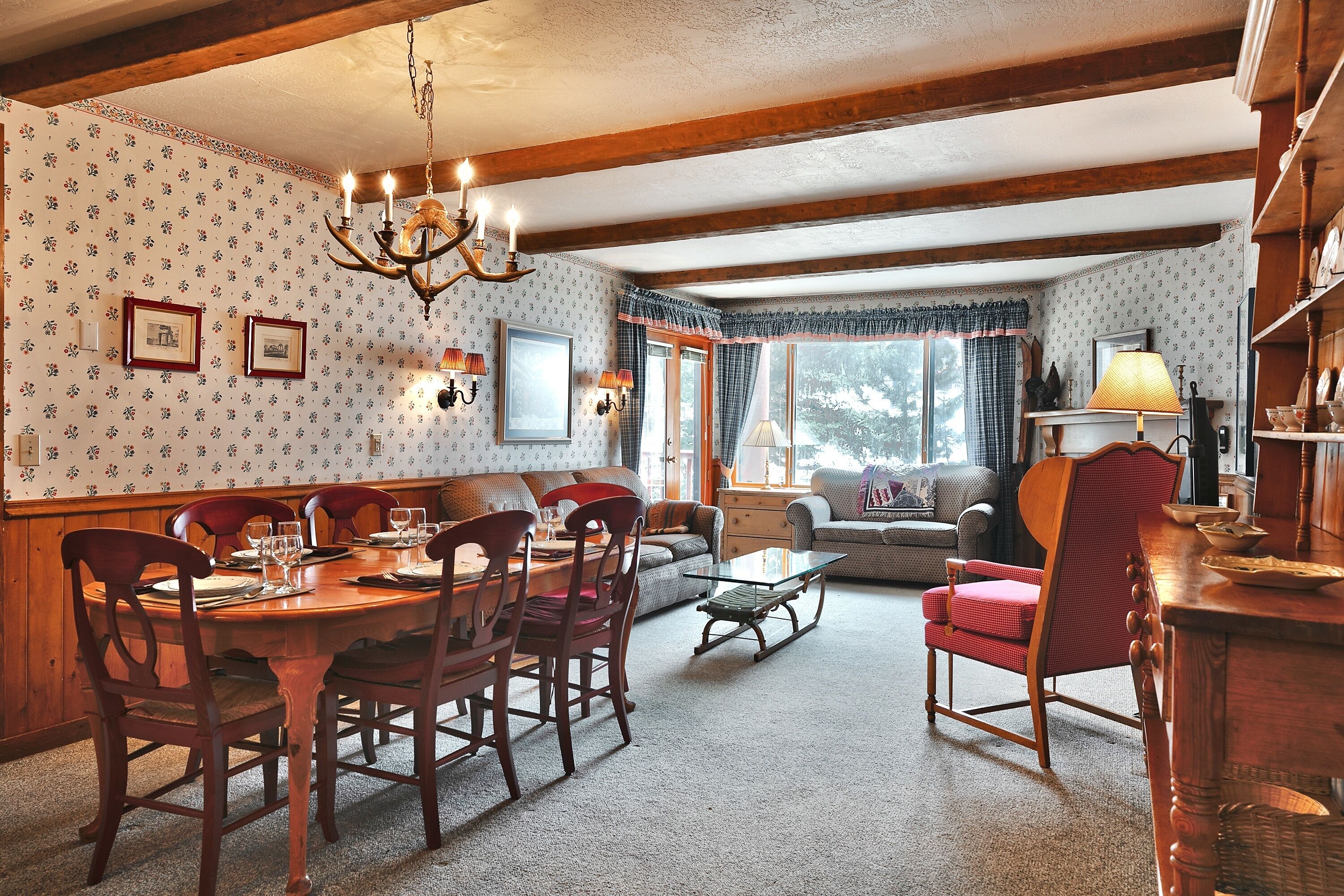 A cozy living room with a wooden dining table, chairs, and a sofa. There are framed pictures on the wall and a chandelier hanging from the ceiling.
