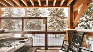 A wooden deck with a hot tub, a bench, and a view of snow-covered trees.