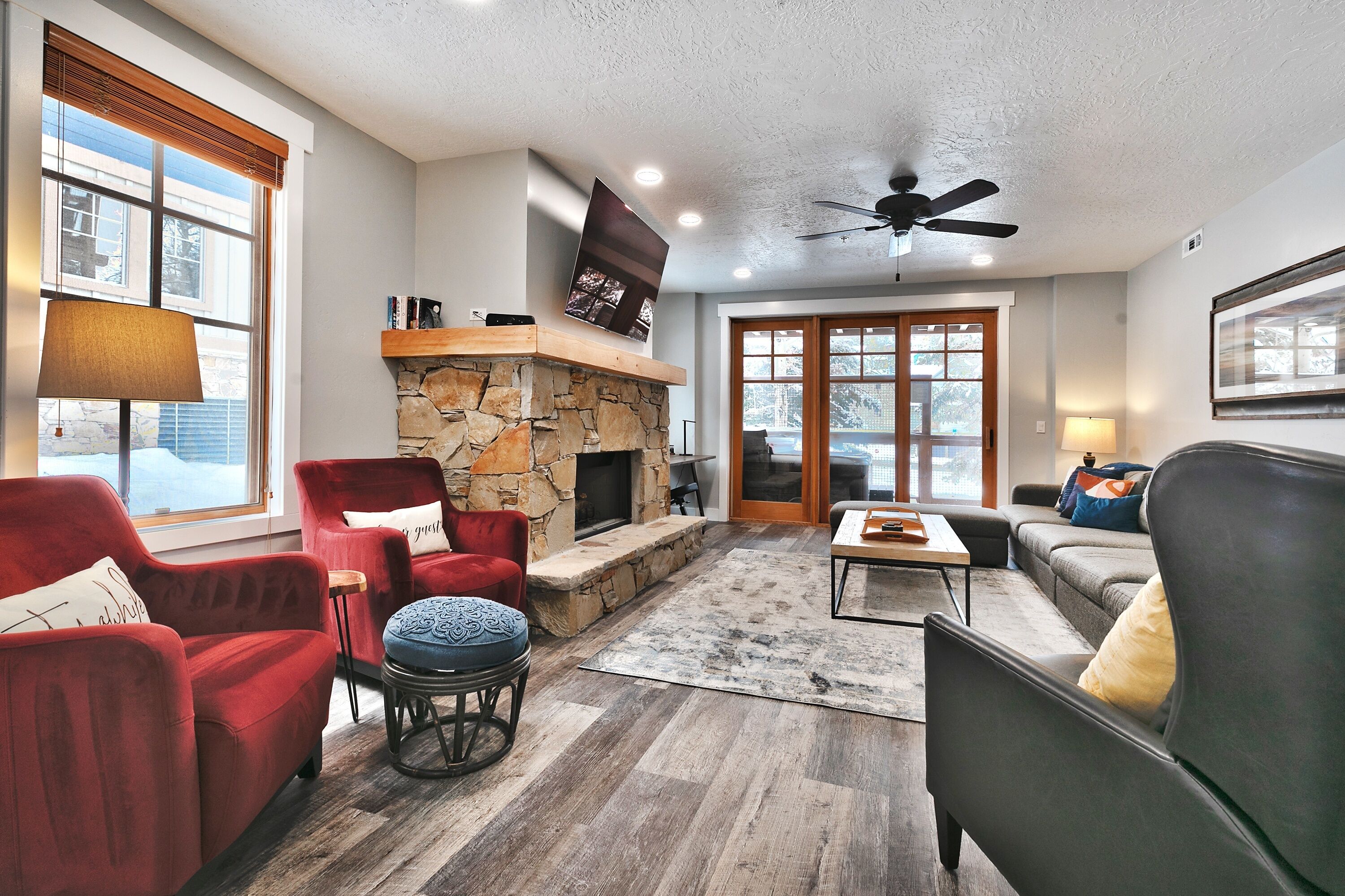 A cozy living room with a stone fireplace, red and gray sofas, a ceiling fan, and a large window.