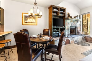 A dining area with a round table, leather chairs, and a chandelier. There is a fireplace with a stone surround and a television in the background.