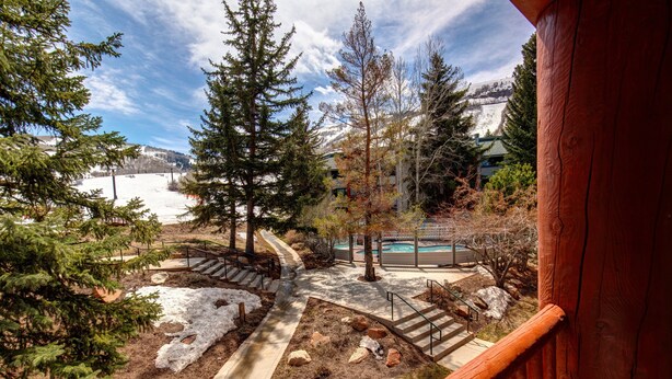 A view from a wooden balcony overlooking a landscaped area with a pool, trees, and a building in the background.