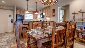 A dining area with a wooden table set for four, surrounded by wooden chairs. A chandelier with antler-like light fixtures hangs above.