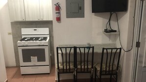 Una cocina pequeña con estufa, una mesa con sillas, extintor, toma de corriente y televisor montado en la pared.