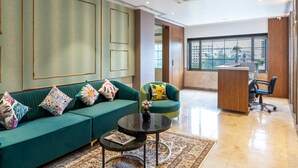 Sala moderna com sofá verde, mesa de centro redonda e poltrona com almofada colorida. Há uma escrivaninha com cadeira e janela com vista para prédios.