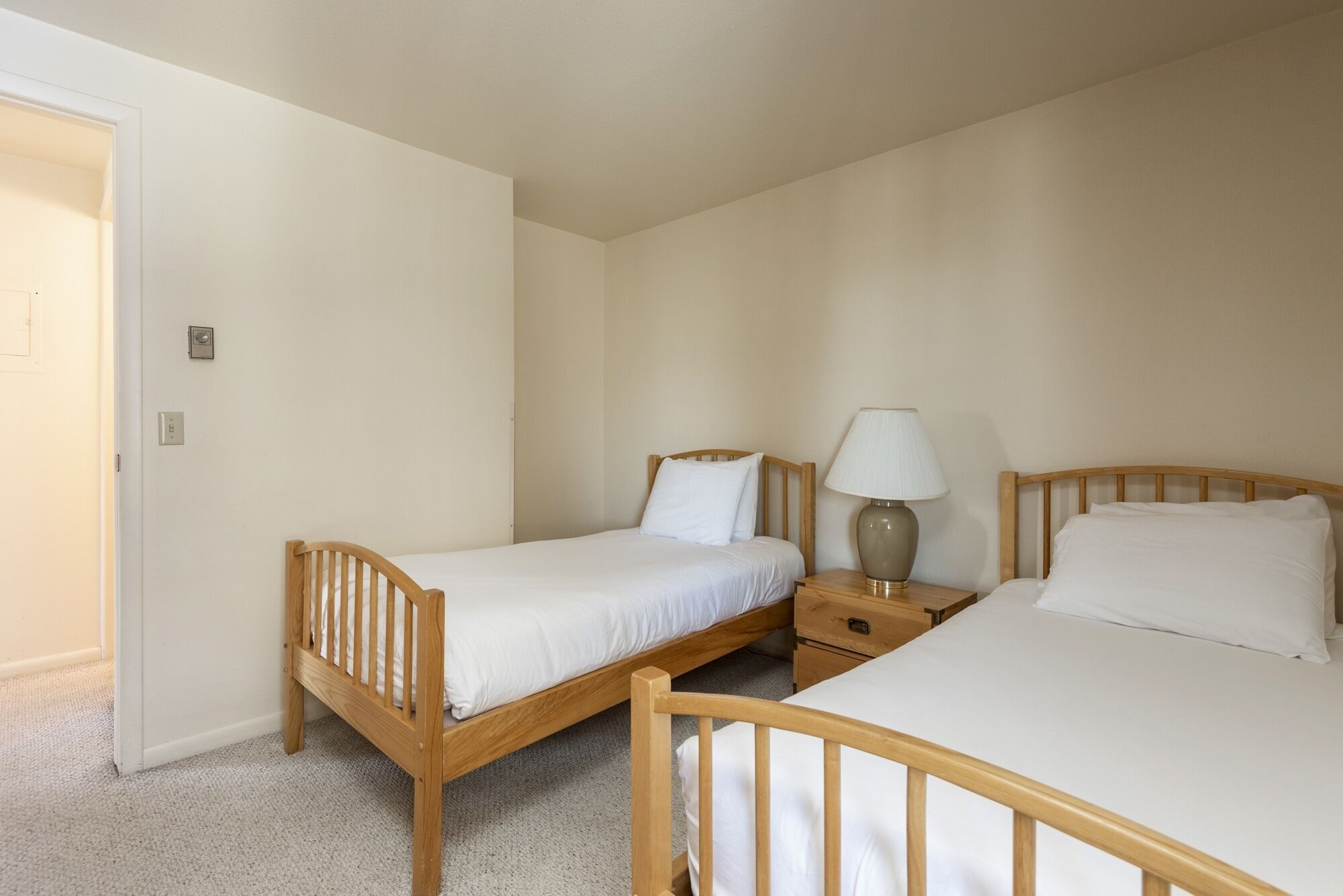 A bedroom with two single beds, a bedside table with a lamp, and a door leading to another room.