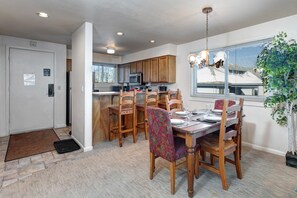 A modern kitchen with wooden cabinets, a dining area with a table set for four, and a large window with blinds. - Snow Flower 141 2 Bedroom Condo by RedAwning (Park City)