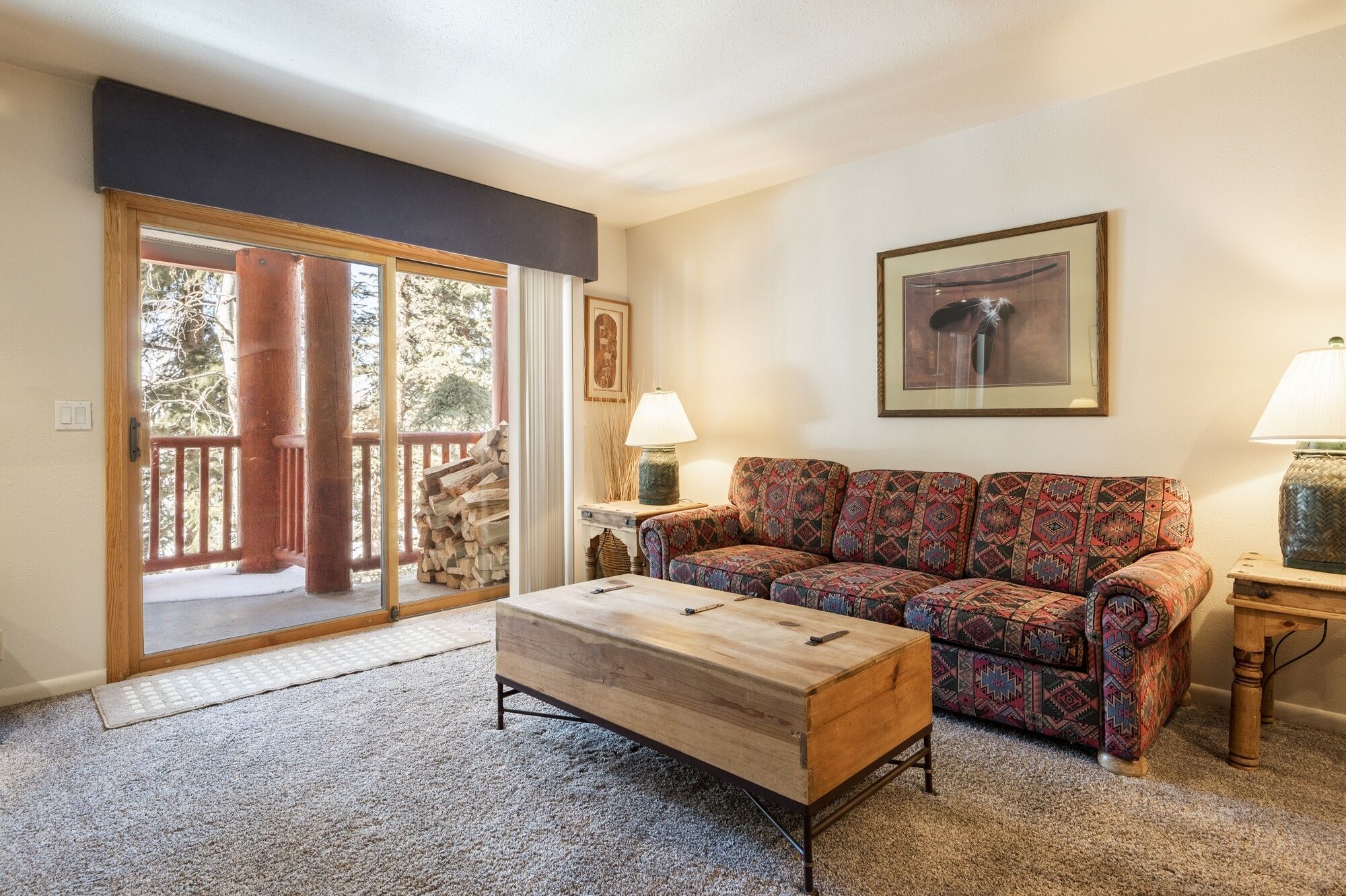 A living room with a sofa, a coffee table, a fireplace, and a door leading to a balcony with a view of trees.