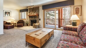 A cozy living room with a stone fireplace, a large wooden coffee table, and a patterned sofa. - Snow Flower 067 2 Bedroom Condo by RedAwning (Park City)