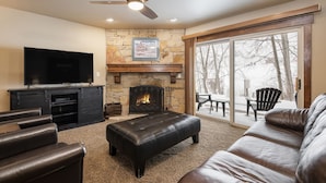 A living room with a stone fireplace, leather furniture, a flat-screen TV, and a ceiling fan. - Snow Flower 109 2 Bedroom Condo by RedAwning (Park City)