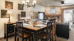 A dining area with a wooden table, chairs, and a chandelier. There is a fireplace with a stone wall behind it and a television on the wall. - Snow Flower 109 2 Bedroom Condo by RedAwning (Park City)
