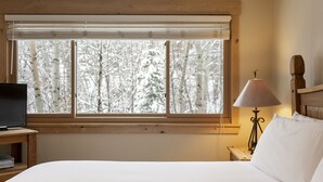 A bedroom with a bed, a TV on a wooden stand, a lamp, and a window with a view of snow-covered trees.