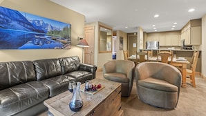 A living room with a black leather sofa, a wooden coffee table with wine glasses, and a painting of a mountain landscape.