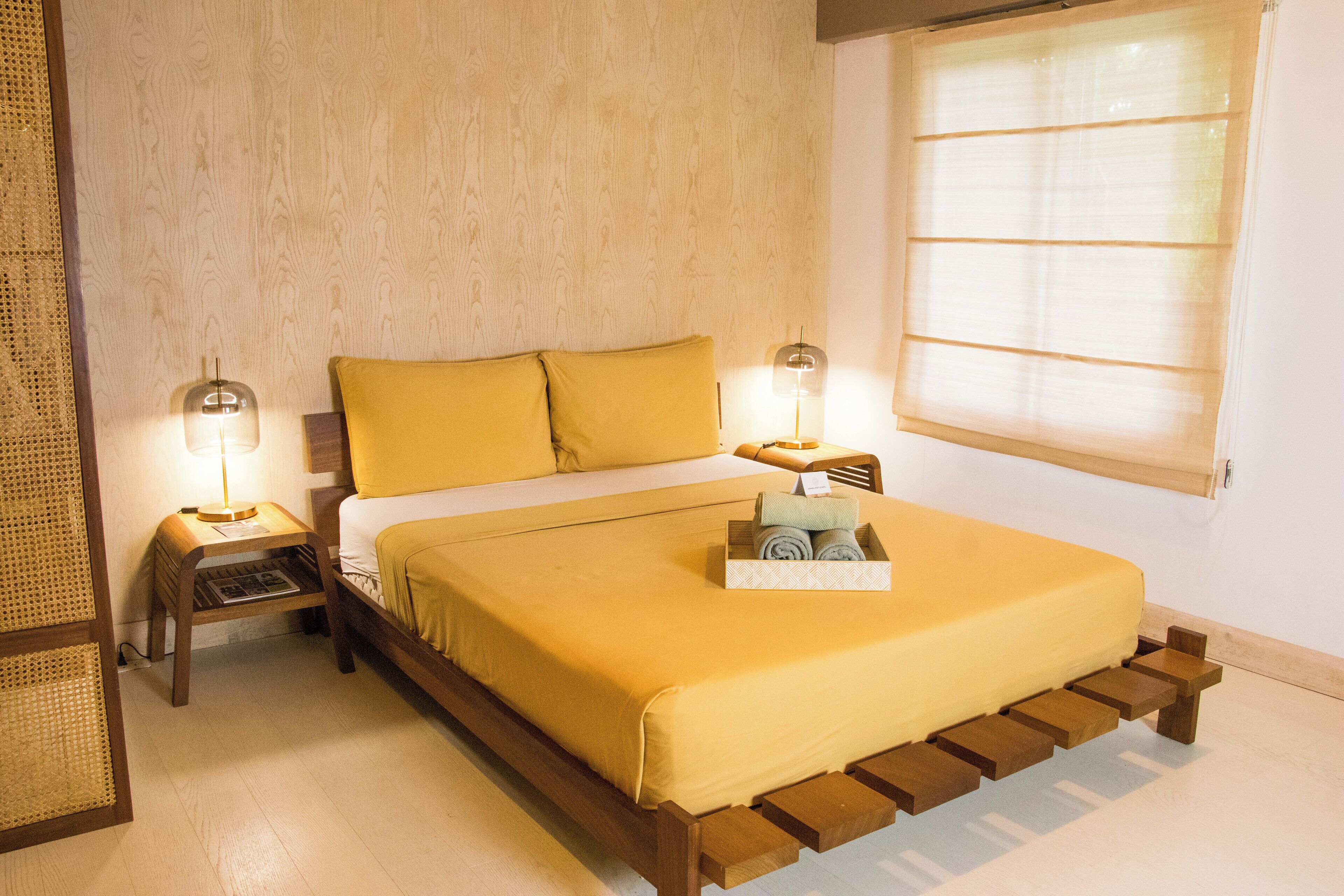 A neatly made bed with a yellow bedspread and pillows, two bedside lamps, a wooden nightstand, and a window with blinds.