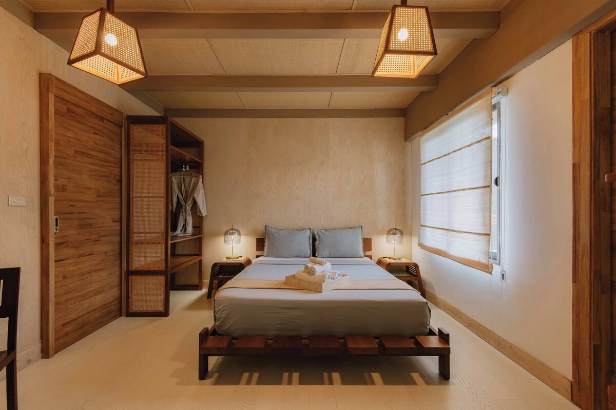 A bedroom with a wooden bed, two bedside tables with lamps, a wardrobe, and a large window with blinds.