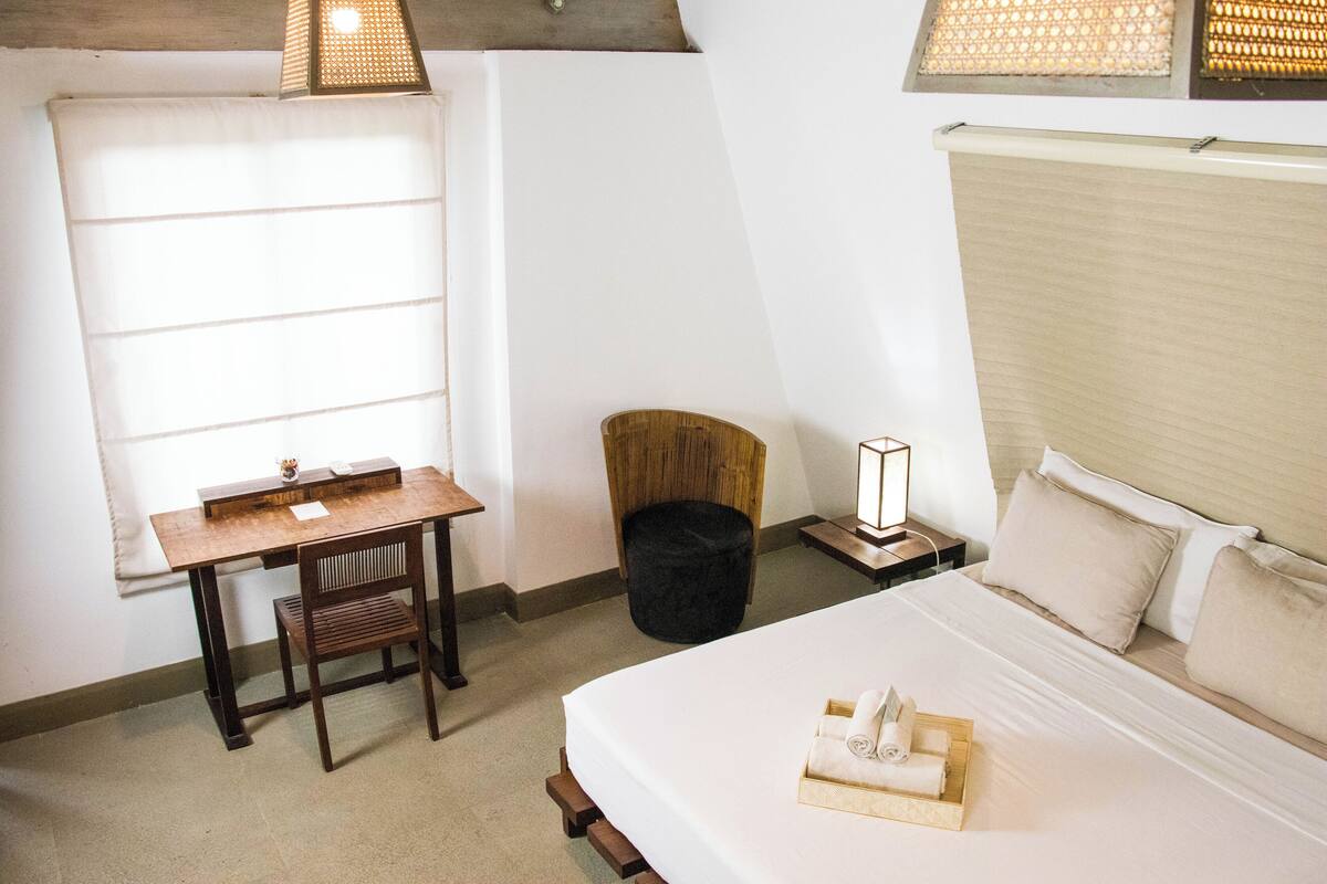 A bedroom with a bed, a desk, a chair, a small stool, and a window with blinds.
