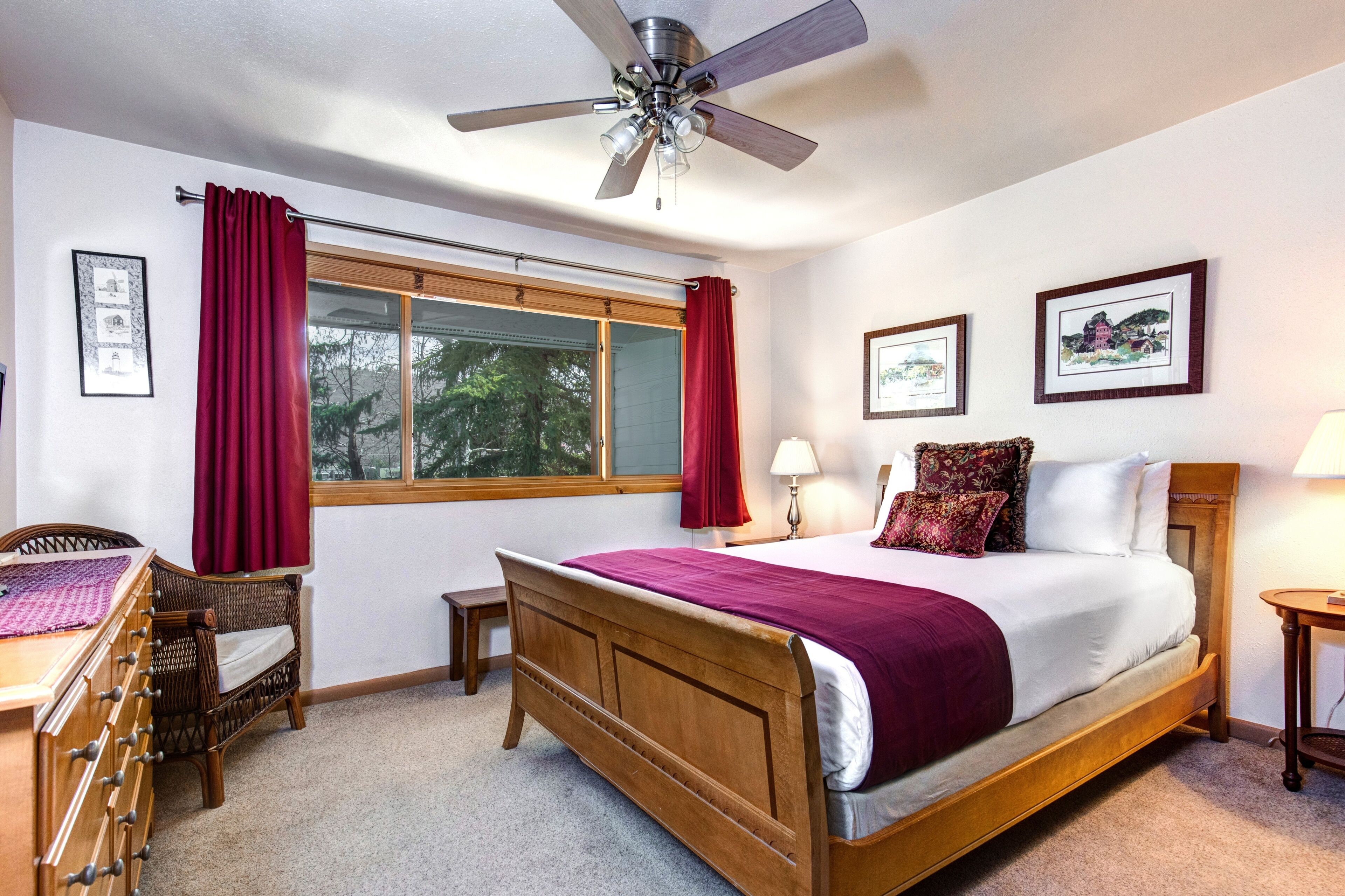 A bedroom with a wooden bed, a ceiling fan, a window with red curtains, a bedside table with a lamp, and framed pictures on the wall.